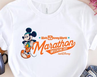 May include: White t-shirt featuring Mickey Mouse in running attire, with the text "Walt Disney World Marathon Weekend 2027 runDisney" in orange. The shirt is paired with blue jeans and a black mug.