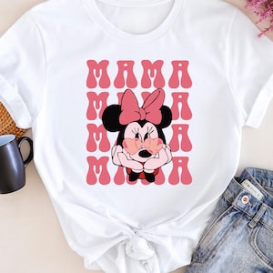 A white t-shirt with a Minnie Mouse graphic and the text "MAMA MAMA" printed on it, along with a pair of jeans and a coffee mug.