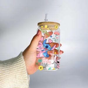 A hand holding a clear glass tumbler with a straw, featuring colorful cartoon characters and flowers printed on it.