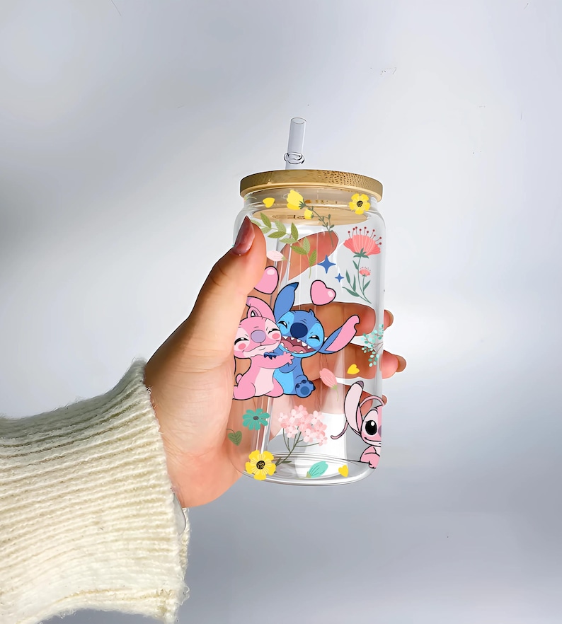 A hand holding a clear glass tumbler with a straw, featuring colorful cartoon characters and flowers printed on it.