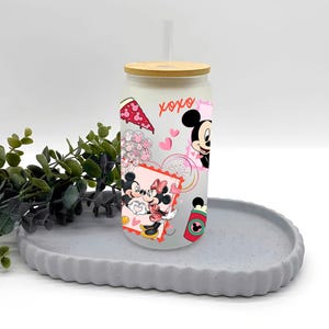 May include: A frosted glass tumbler with a bamboo lid and a clear straw. The tumbler is decorated with cartoon images of Mickey and Minnie Mouse, hearts, and the word "xoxo". The tumbler is sitting on a gray, round tray.