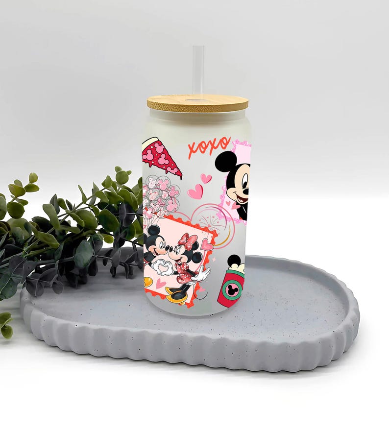 May include: A frosted glass tumbler with a bamboo lid and a clear straw. The tumbler is decorated with cartoon images of Mickey and Minnie Mouse, hearts, and the word "xoxo". The tumbler is sitting on a gray, round tray.