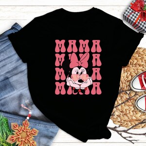 a black t-shirt with a pink Minnie Mouse graphic and the text "MAMA MINNIE" printed on it. The t-shirt is placed on a wooden surface, surrounded by various items such as a pair of jeans, a gift box, and a pair of red and white sneakers.