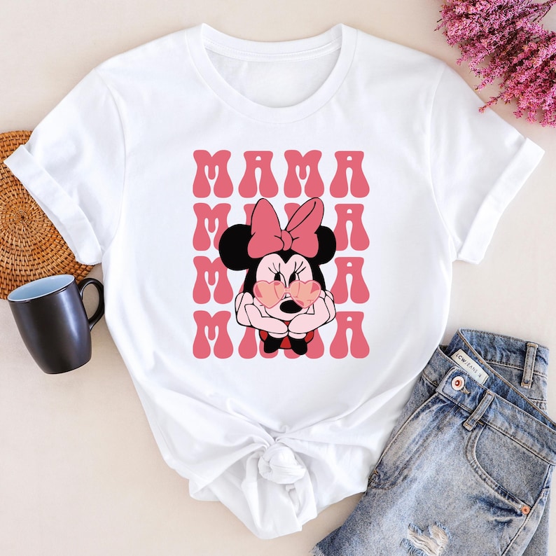 A white t-shirt with a Minnie Mouse graphic and the text "MAMA MAMA" printed on it, along with a pair of jeans and a coffee mug.