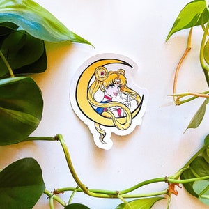 May include: A sticker featuring a cartoon illustration of a woman with blonde hair and a crescent moon behind her. She is wearing a blue sailor uniform and holding a pair of glasses. The sticker is on a white background.