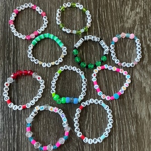 May include: A collection of 11 colourful beaded bracelets with different words spelled out on them. The words include "Cause We Were In Paris", "Home To My Cats", "Cuz She's Dead Ready", "Wendy For The End", "I've Got The Goodie", "Skies", "Red Lip Classic", "Peter Is My Life", "Losing", "16th Avenue", "Cherry Lips Crystal".