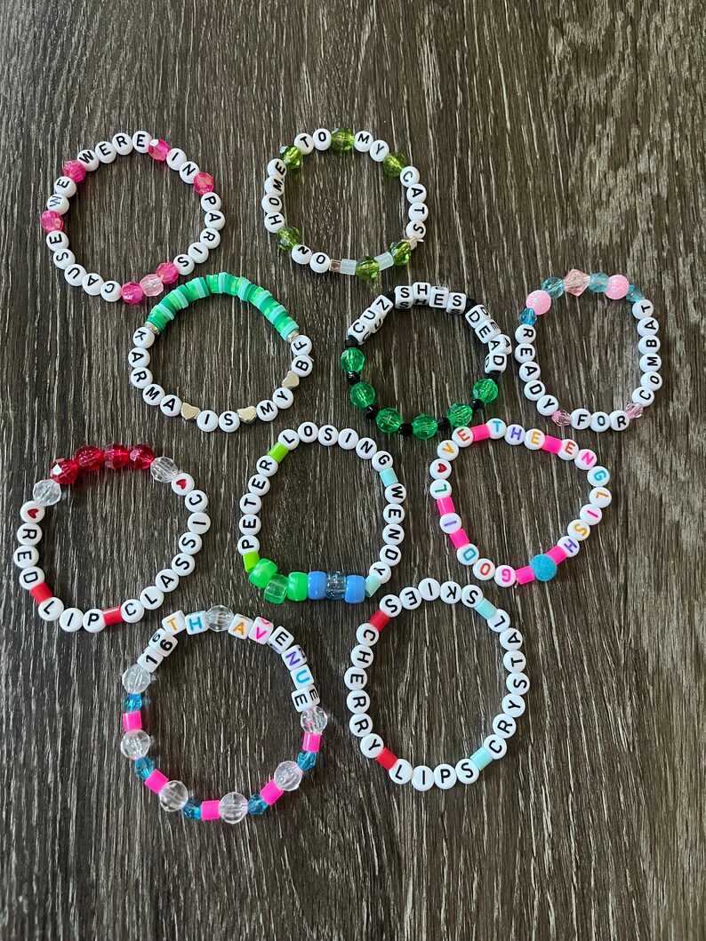 May include: A collection of 11 colourful beaded bracelets with different words spelled out on them. The words include "Cause We Were In Paris", "Home To My Cats", "Cuz She's Dead Ready", "Wendy For The End", "I've Got The Goodie", "Skies", "Red Lip Classic", "Peter Is My Life", "Losing", "16th Avenue", "Cherry Lips Crystal".