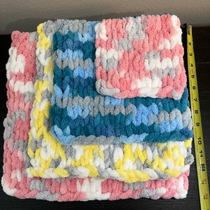 May include: Four crocheted blankets in different colors and patterns, stacked on top of each other. The blankets are made with a chunky yarn and have a soft, fluffy texture. The colors are pink, yellow, blue, and gray.