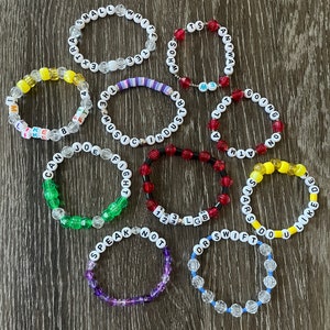 May include: A collection of 12 colourful beaded bracelets with various words and phrases spelled out on them. The bracelets are made with clear, yellow, red, green, purple, and blue beads.