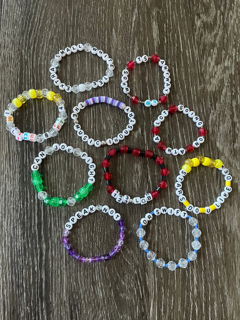 May include: A collection of 12 colourful beaded bracelets with various words and phrases spelled out on them. The bracelets are made with clear, yellow, red, green, purple, and blue beads.