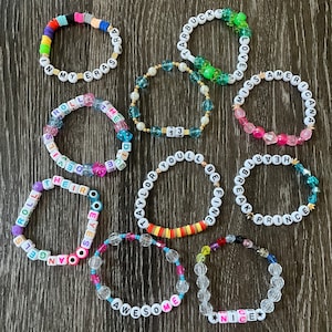 May include: A collection of 11 colourful beaded bracelets with various words and phrases, including "In My Eras", "Roll With The Devil", "Taylor You'll Be Fine", "Awesome", "Nice", "Starbucks", "Lover", "Miss Americana", "Hear Break Prince", and "Their Eyes", on a wooden background.