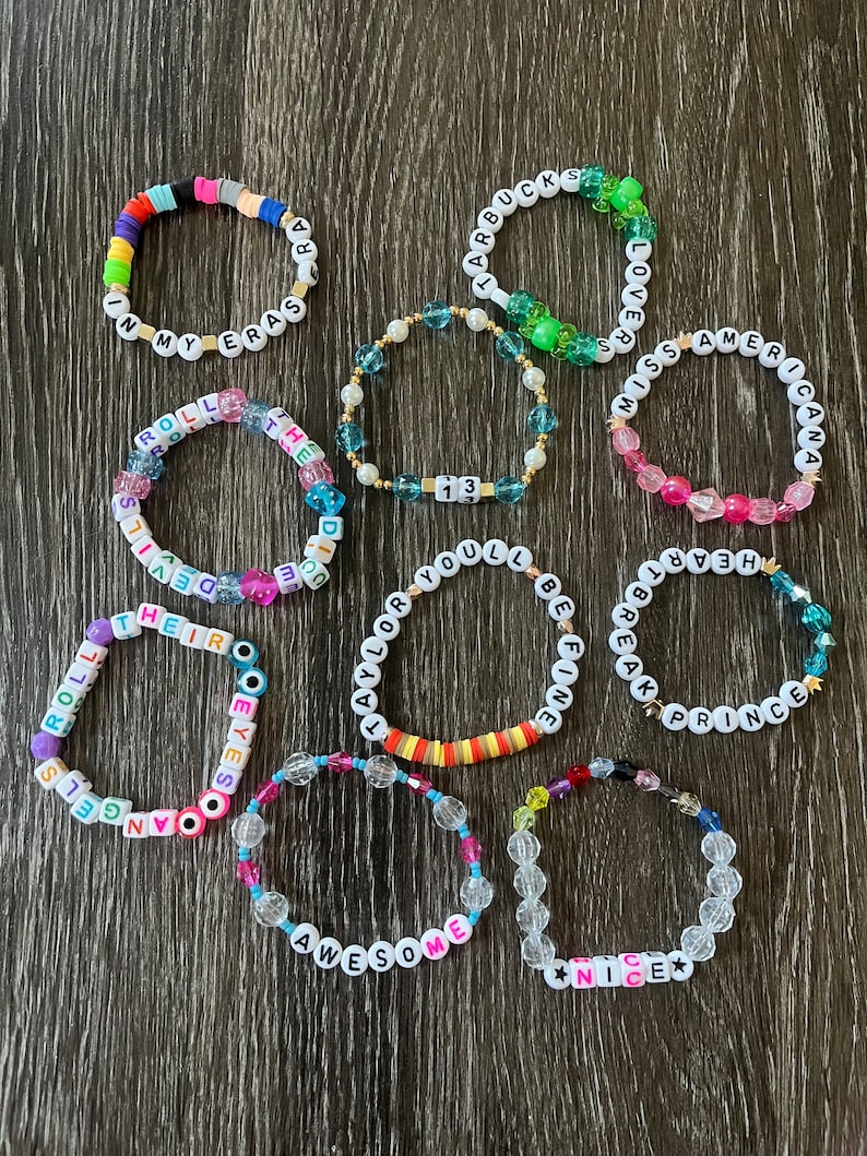 May include: A collection of 11 colourful beaded bracelets with various words and phrases, including "In My Eras", "Roll With The Devil", "Taylor You'll Be Fine", "Awesome", "Nice", "Starbucks", "Lover", "Miss Americana", "Hear Break Prince", and "Their Eyes", on a wooden background.