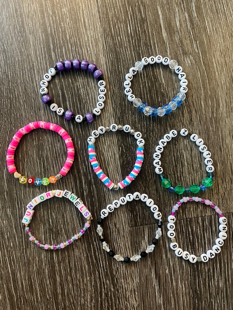 May include: Ten colourful beaded bracelets with different song titles and lyrics from the singer Taylor Swift. The bracelets are made with white letter beads and colourful beads.