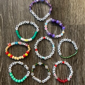 May include: Nine colourful beaded bracelets with different words spelled out on them. The bracelets are made with various colours of beads, including red, green, blue, purple, and white.