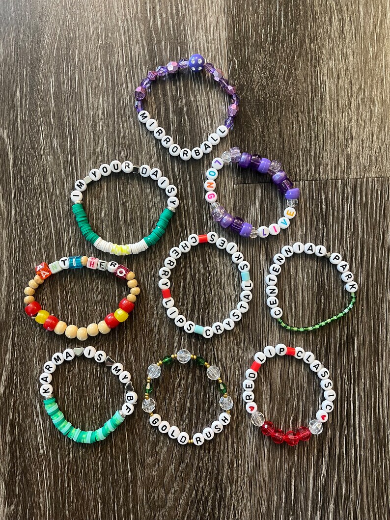 May include: Nine colourful beaded bracelets with different words spelled out on them. The bracelets are made with various colours of beads, including red, green, blue, purple, and white.