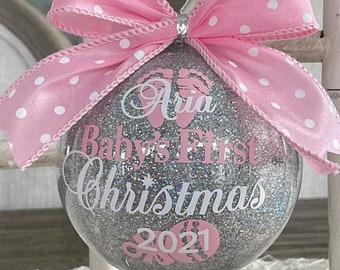 Può includere: Un addobbo natalizio in glitter argento con un fiocco rosa e il testo "Aria Baby's First Christmas 2021".