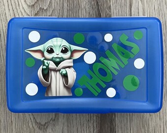 May include: A blue lunchbox featuring a cartoon image of a green alien character with large ears and a white robe. The name "THOMAS" is printed in green, along with white and green polka dots.