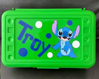 May include: A bright green lunchbox featuring the name "Troy" in blue, along with a cartoon image of Stitch from Lilo & Stitch. The lunchbox has a green lid and is decorated with white and blue polka dots.