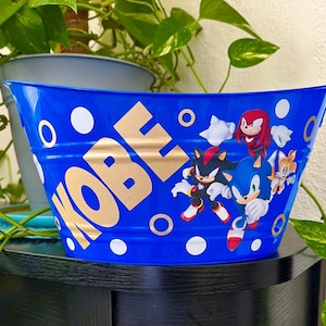 May include: A blue metal bucket with the word "KOBE" in gold lettering. The bucket features cartoon characters and white circles. The bucket is sitting on a black surface.
