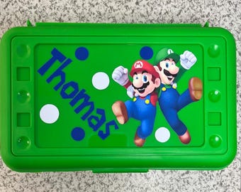 May include: Green plastic pencil case with a Mario and Luigi design. The case has a white background with blue polka dots and the name "Thomas" written in blue letters.