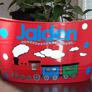 May include: Red metal bucket with a train design. The bucket has the name "Jaiden" in blue letters. The train is red, blue, and green with white smoke and clouds. White and blue polka dots decorate the sides.