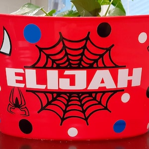 May include: Red metal bucket with a black spider web design, black spider silhouettes, and the name "ELIJAH" in white letters. The bucket is decorated with black and white polka dots and blue and white circles.