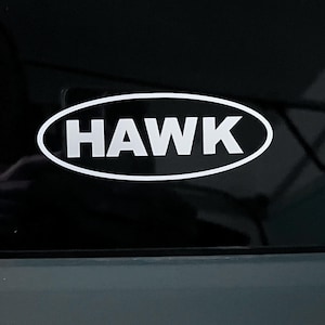 May include: A white oval decal featuring the word HAWK in bold, capitalized, sans-serif lettering. The design consists of a white outline forming an oval shape with the text HAWK centered inside, creating a high-contrast graphic element.