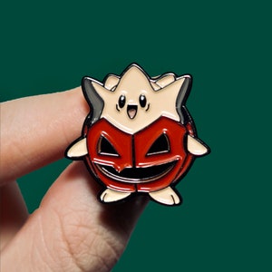 May include: An enamel pin featuring a cartoon character dressed as a jack-o'-lantern. The character has a star-shaped head and a wide smile.