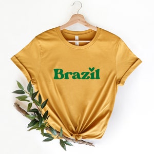 May include: A mustard yellow t-shirt with a green graphic that says "Brazil" with a small heart above the "z".