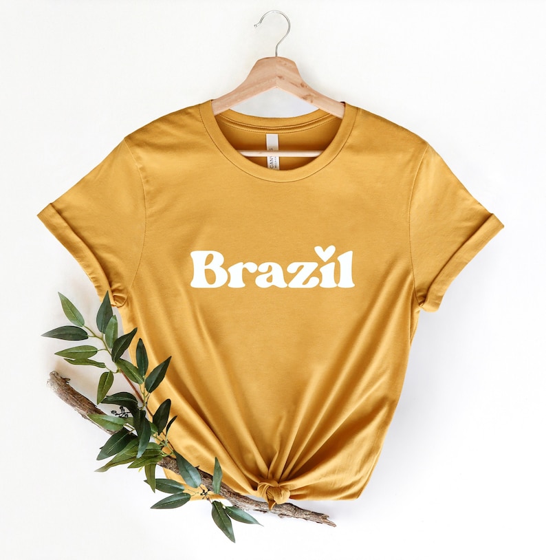 May include: A mustard yellow t-shirt with a white graphic that says "Brazil" with a small heart after the "l".