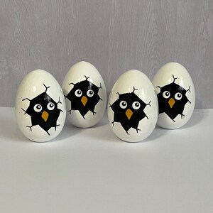 May include: Four white eggs with black and yellow painted bird faces peeking out of the cracked shell.