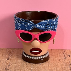 May include: A decorative terracotta flower pot painted to resemble a face. It features a blue bandana, pink sunglasses, a pearl necklace, and gold hoop earrings. The lips are painted red, and the nose is a small beige sphere.