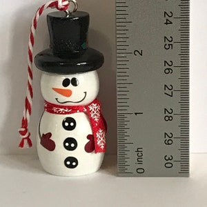 May include: A white snowman ornament with a black top hat, red mittens, and a red scarf with white snowflakes. The snowman has three black buttons and a carrot nose.