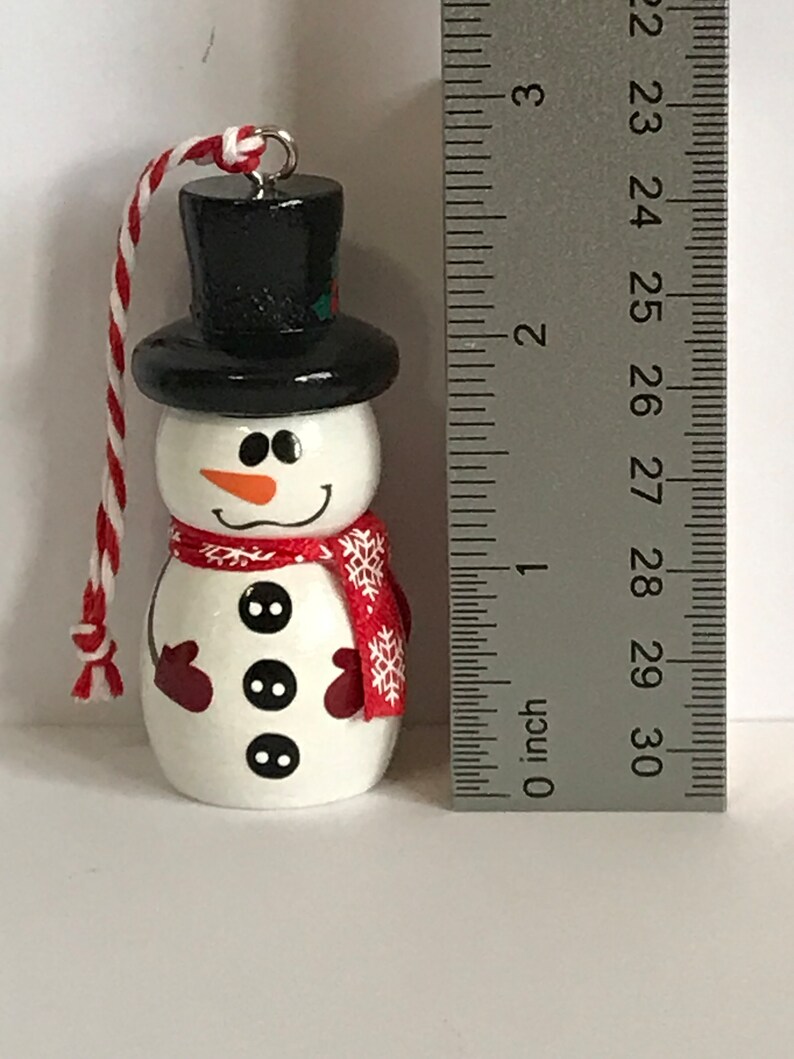 May include: A white snowman ornament with a black top hat, red mittens, and a red scarf with white snowflakes. The snowman has three black buttons and a carrot nose.