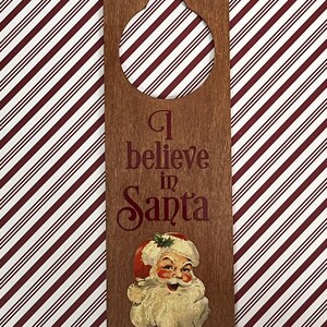 May include: Wooden door hanger with a round cutout for a door knob. The hanger is brown and has the text "I believe in Santa" printed in red. A vintage image of Santa Claus is printed on the bottom of the hanger.