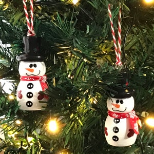 May include: Two white snowman Christmas ornaments with black hats, red scarves, and black buttons. The ornaments are hanging on a green Christmas tree with white lights.
