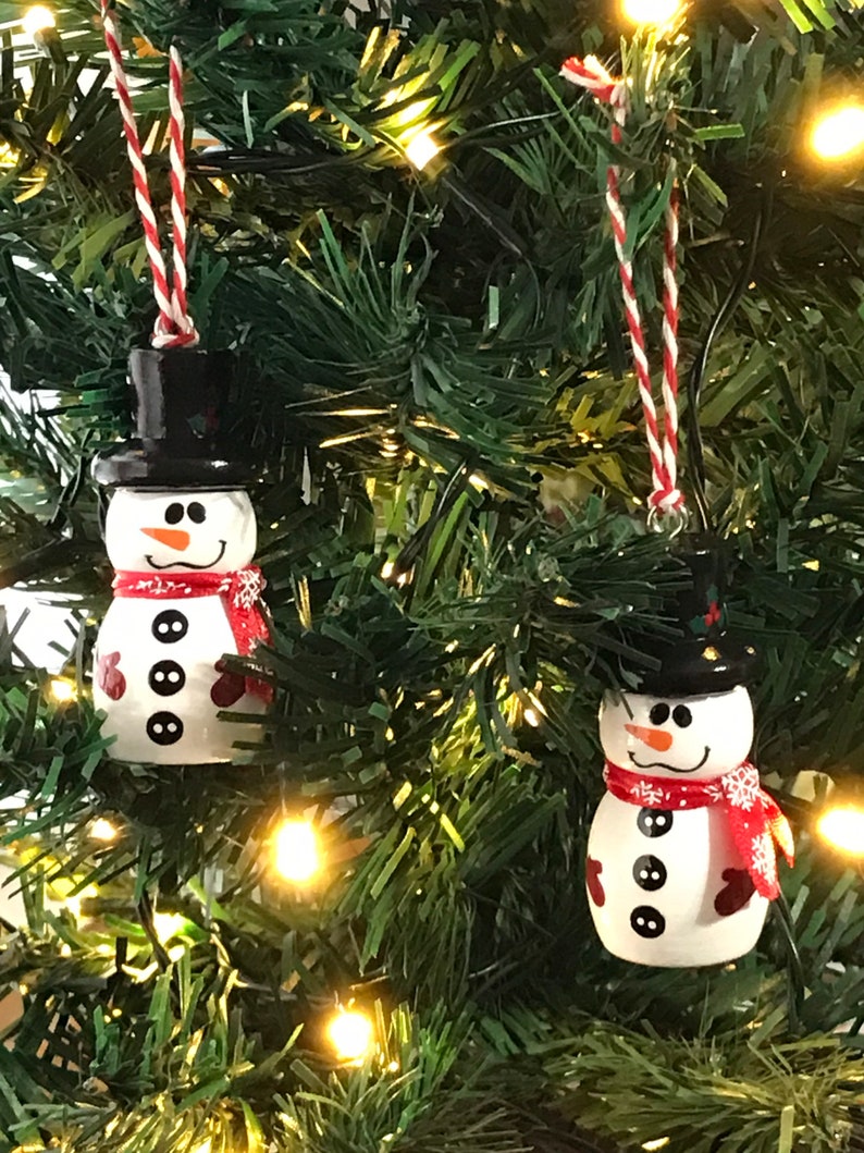 May include: Two white snowman Christmas ornaments with black hats, red scarves, and black buttons. The ornaments are hanging on a green Christmas tree with white lights.