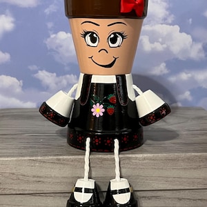 May include: A hand-painted terracotta flower pot shaped like a doll. The doll has a brown top, a red bow, and a black dress with a strawberry and flower design. The doll's arms are white cups, and its legs are black shoes.