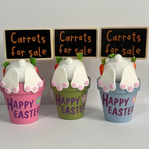 May include: Three ceramic bunny planter pots with carrot tops and white bunny tails sticking out. The pots are painted in pink, green, and blue with the words "Happy Easter" on the front. Each pot has a small wooden sign that reads "Carrots for sale".