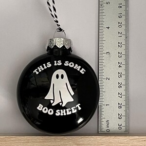 May include: Black round Christmas ornament with the text "THIS IS SOME BOO SHEET" and a white ghost illustration. The ornament has a silver cap and a black and white string for hanging. A ruler is next to the ornament.