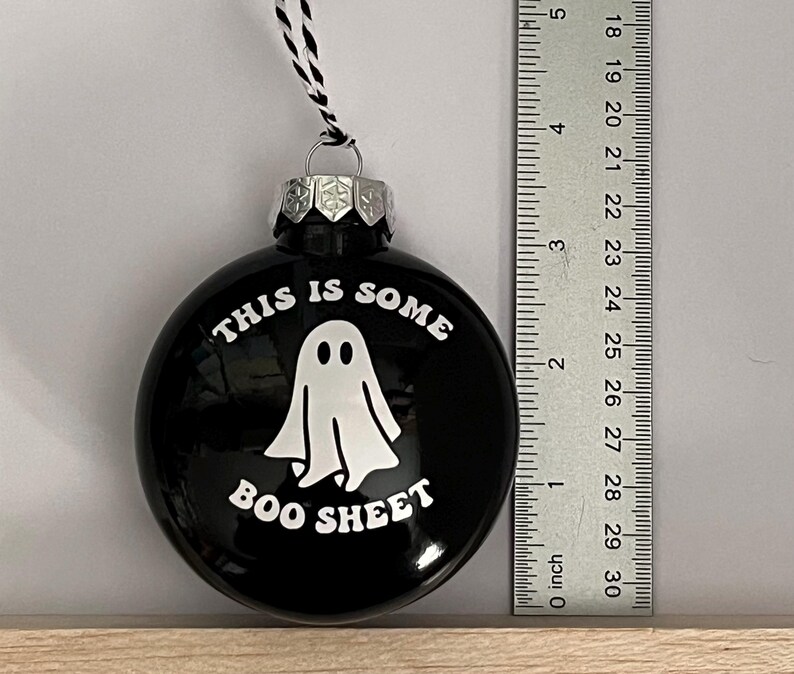 May include: Black round Christmas ornament with the text "THIS IS SOME BOO SHEET" and a white ghost illustration. The ornament has a silver cap and a black and white string for hanging. A ruler is next to the ornament.