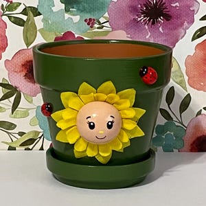 May include: A green painted terracotta flower pot with a matching saucer. The pot is decorated with a yellow sunflower and two red ladybugs. The sunflower has a smiling face. The pot is designed for planting flowers.
