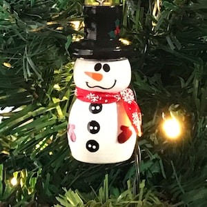 May include: A white snowman Christmas ornament with a black top hat, red scarf with white snowflakes, and black buttons. The snowman has a carrot nose and a black mouth.