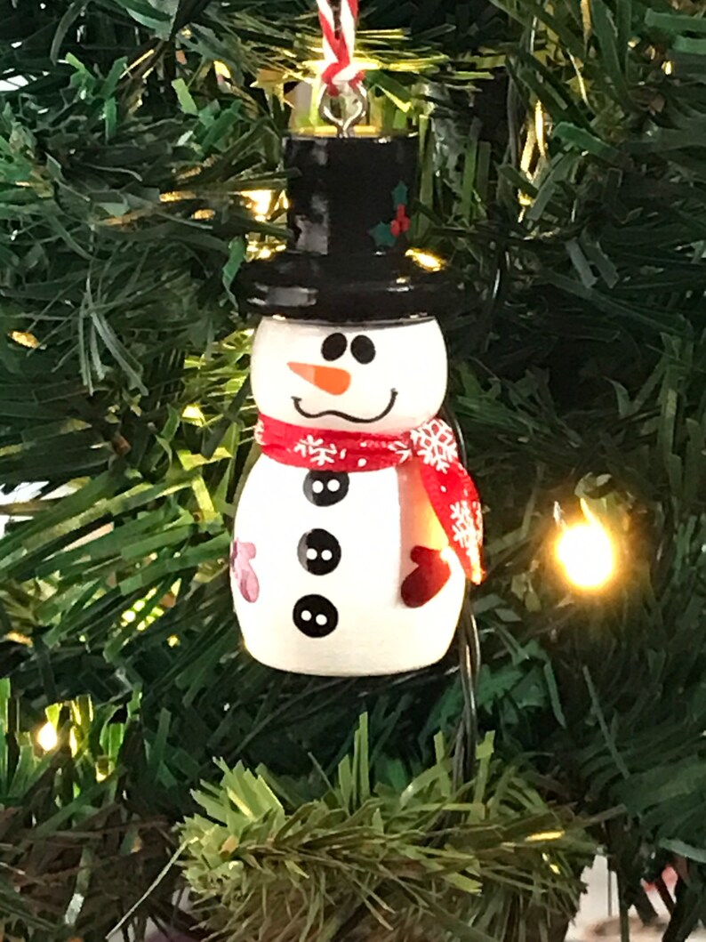 May include: A white snowman Christmas ornament with a black top hat, red scarf with white snowflakes, and black buttons. The snowman has a carrot nose and a black mouth.