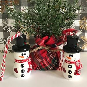 May include: Two white snowman ornaments with black hats, red scarves, and black buttons. The ornaments are hanging from red and white twine. The snowmen are in front of a plaid fabric bag and a green bush.