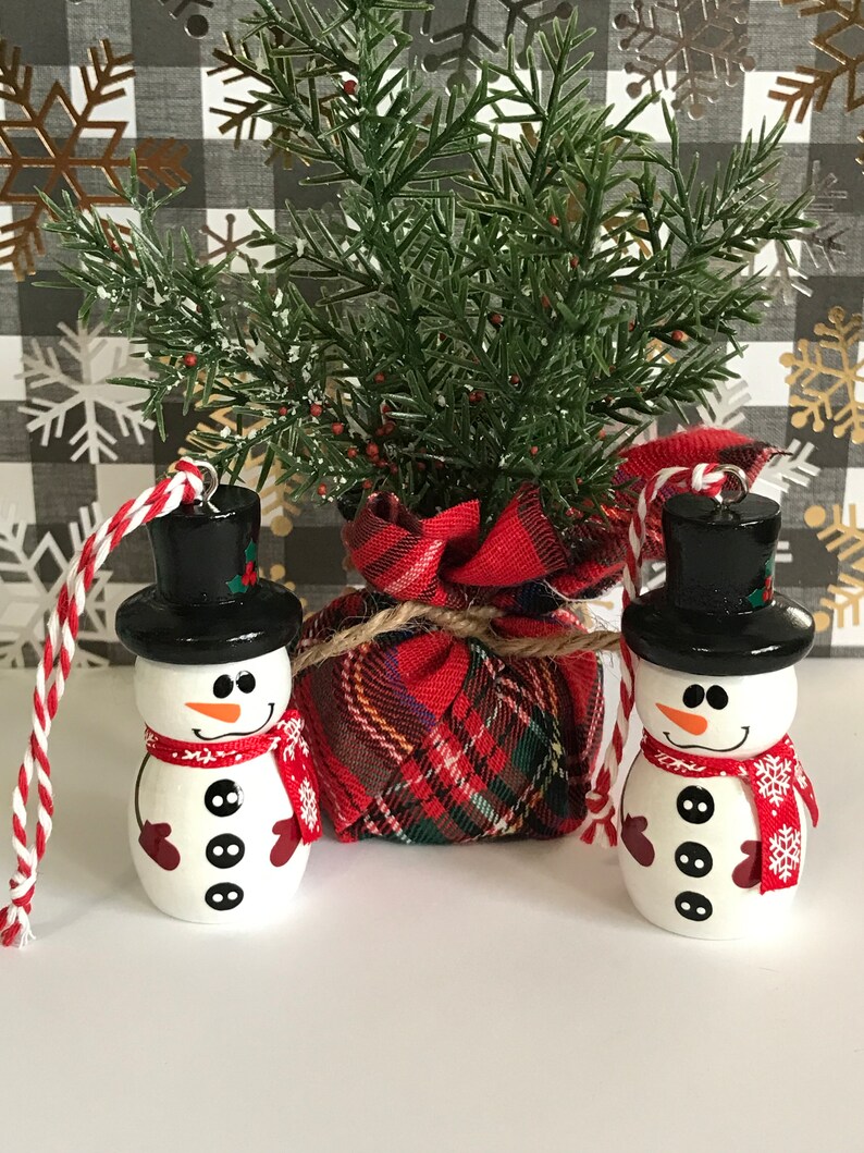 May include: Two white snowman ornaments with black hats, red scarves, and black buttons. The ornaments are hanging from red and white twine. The snowmen are in front of a plaid fabric bag and a green bush.