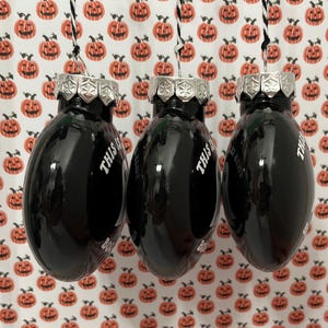 May include: Three black oval-shaped Christmas ornaments with silver tops and the text "THIS IS BOO SHEET" in white. The ornaments hang from black and white string against a background of orange pumpkin-patterned paper.
