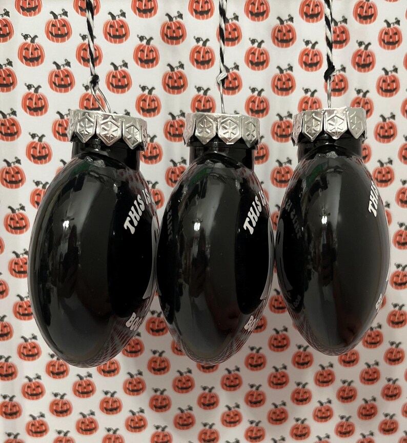 May include: Three black oval-shaped Christmas ornaments with silver tops and the text "THIS IS BOO SHEET" in white. The ornaments hang from black and white string against a background of orange pumpkin-patterned paper.