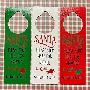 May include: Three wooden door hangers with a red, white, and green background. Each hanger has a cut-out for a door knob and says "Santa Please Stop Here For [Name] We Have Cookies". The names are Presley, Natalie, and Amanda.