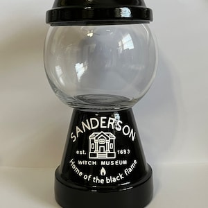 May include: Black and white glass candy jar with a black lid and a white knob. The jar has the words "SANDERSON est. 1693 WITCH MUSEUM Home of the black flame" written on it.